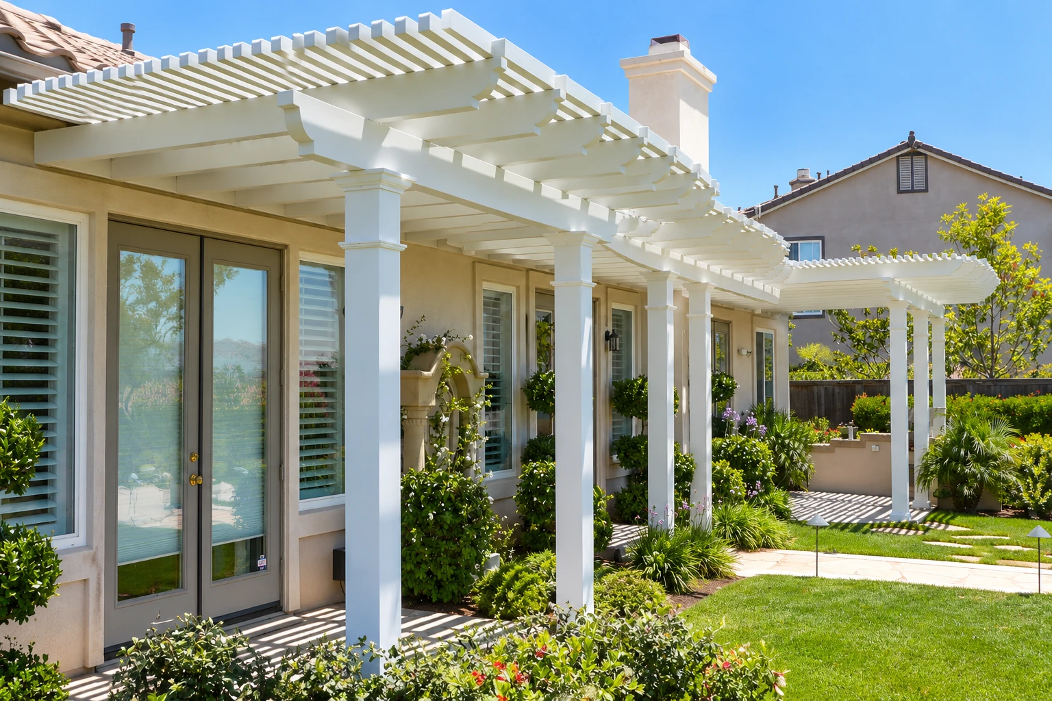 A white patio cover with evenly spaced slats extends over a house-side walkway, supported by square columns amid landscaped gardens.