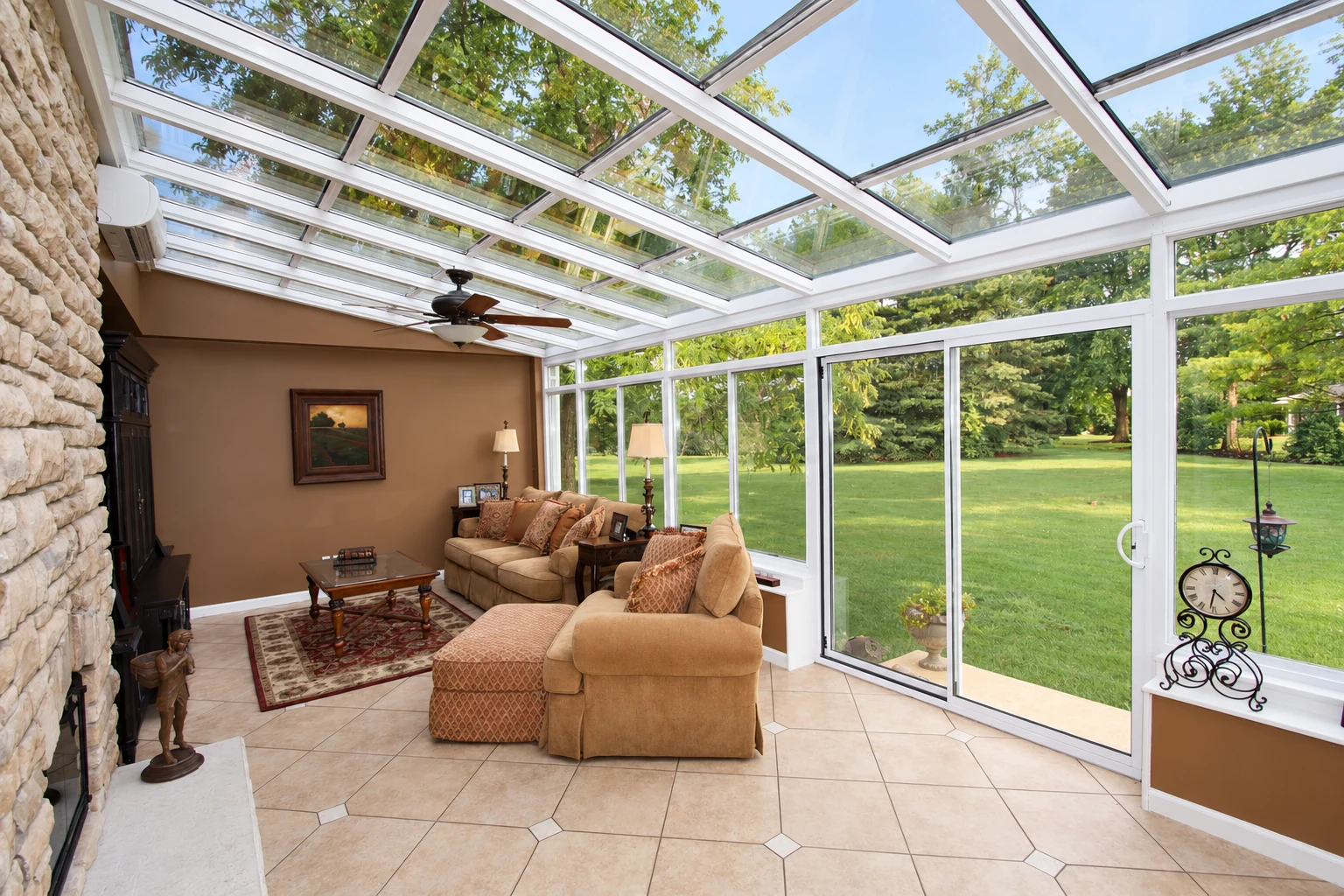 Glass-roofed solarium furnished with tan sofas and a tiled floor, overlooking a green lawn.