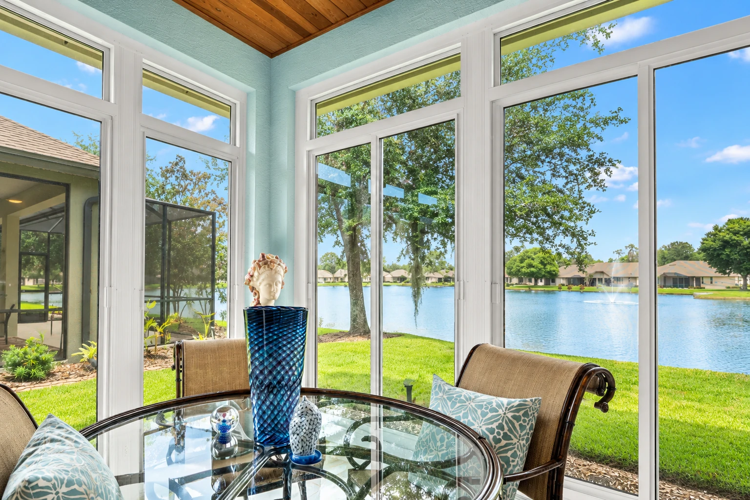 Sunroom addition with white-framed windows overlooking a lake, furnished with a glass table and wicker chairs.