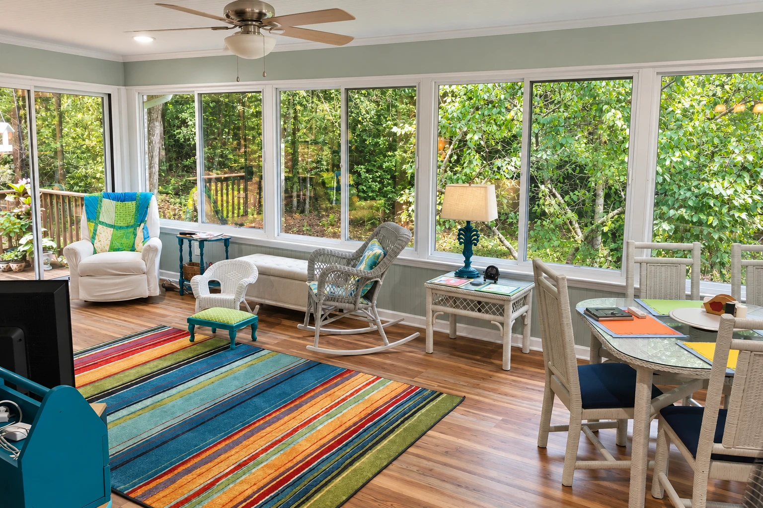 Sunroom with broad windows overlooking trees, wicker seating, a colorful striped rug, and a dining table.
