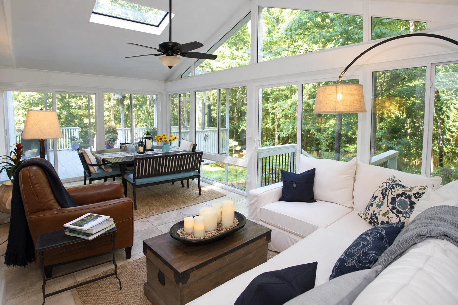 A bright sunroom with floor-to-ceiling windows, a white sofa, a dining table, and views of leafy trees.